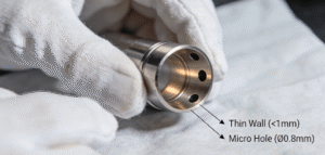 Close-up of a machined titanium medical component with challenging features: an ultra-thin wall section and several deep micro-holes, held by gloved hands.
